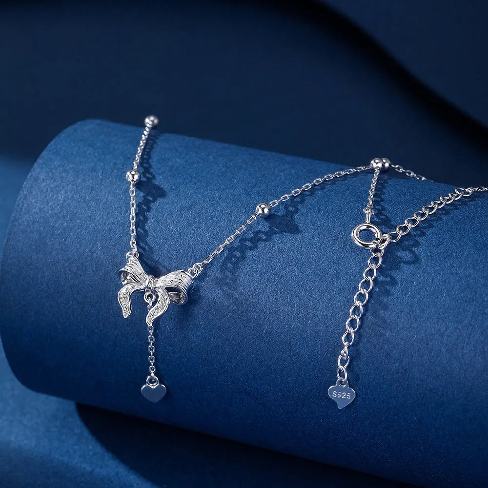 A close-up of a silver necklace with a heart and bow pendant on an O-ring chain.