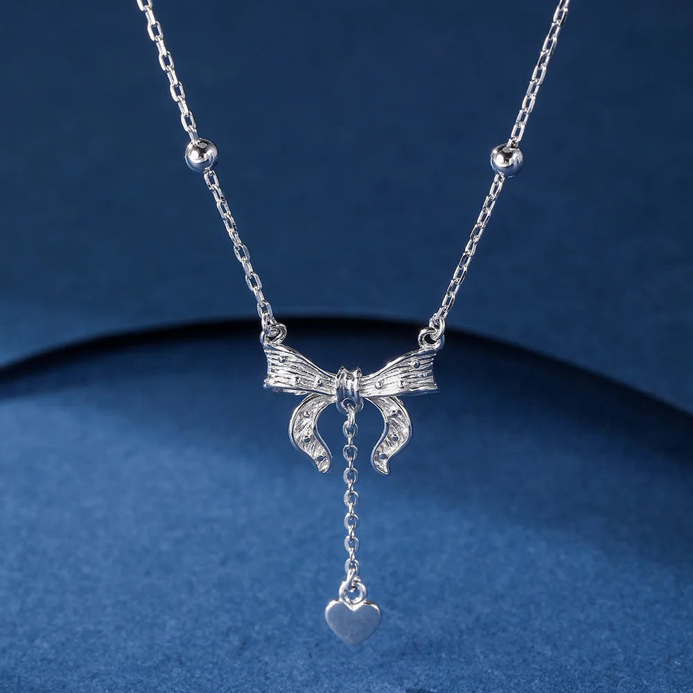 A close-up of a silver necklace with a heart and bow pendant on an O-ring chain.