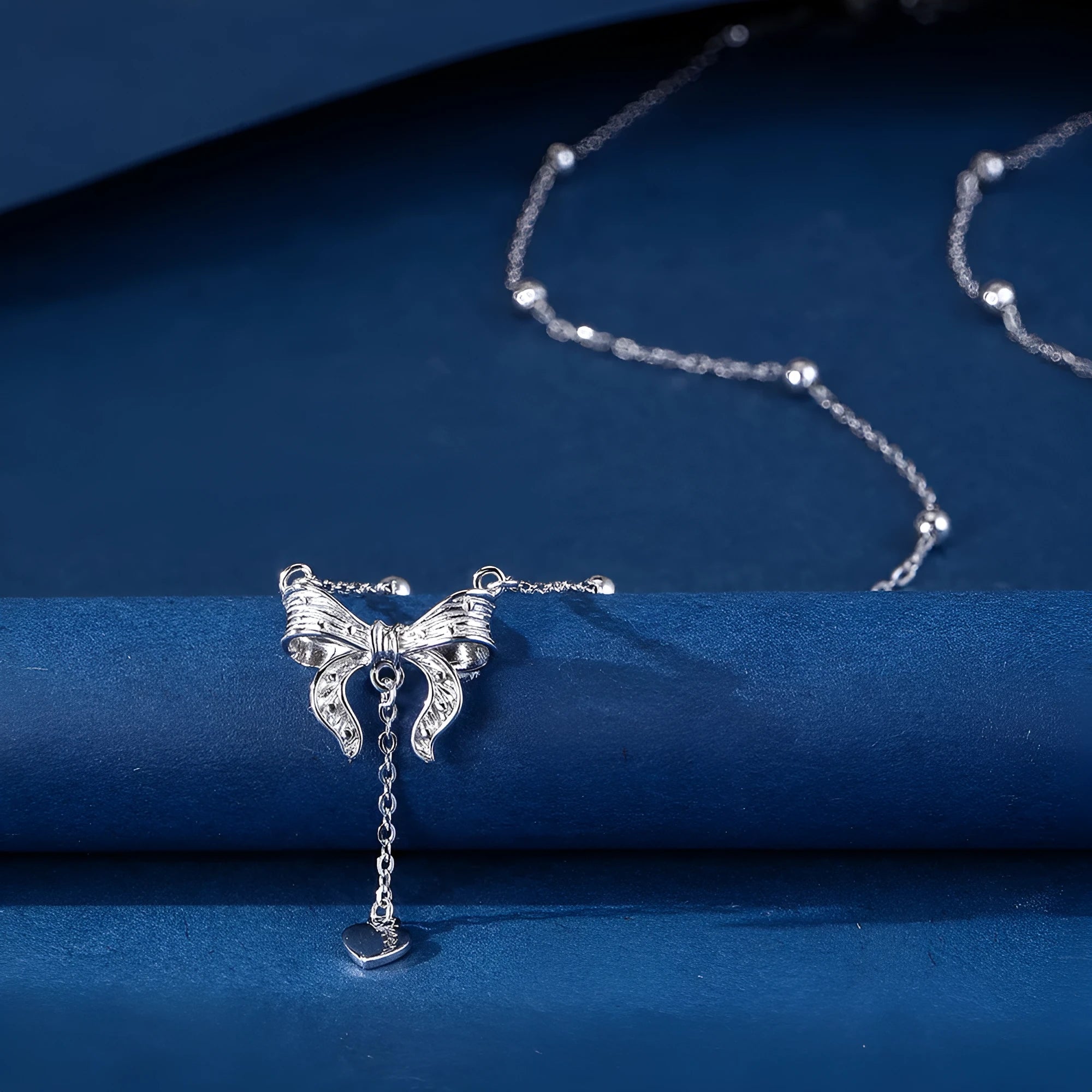 A close-up of a silver necklace with a heart and bow pendant on an O-ring chain.