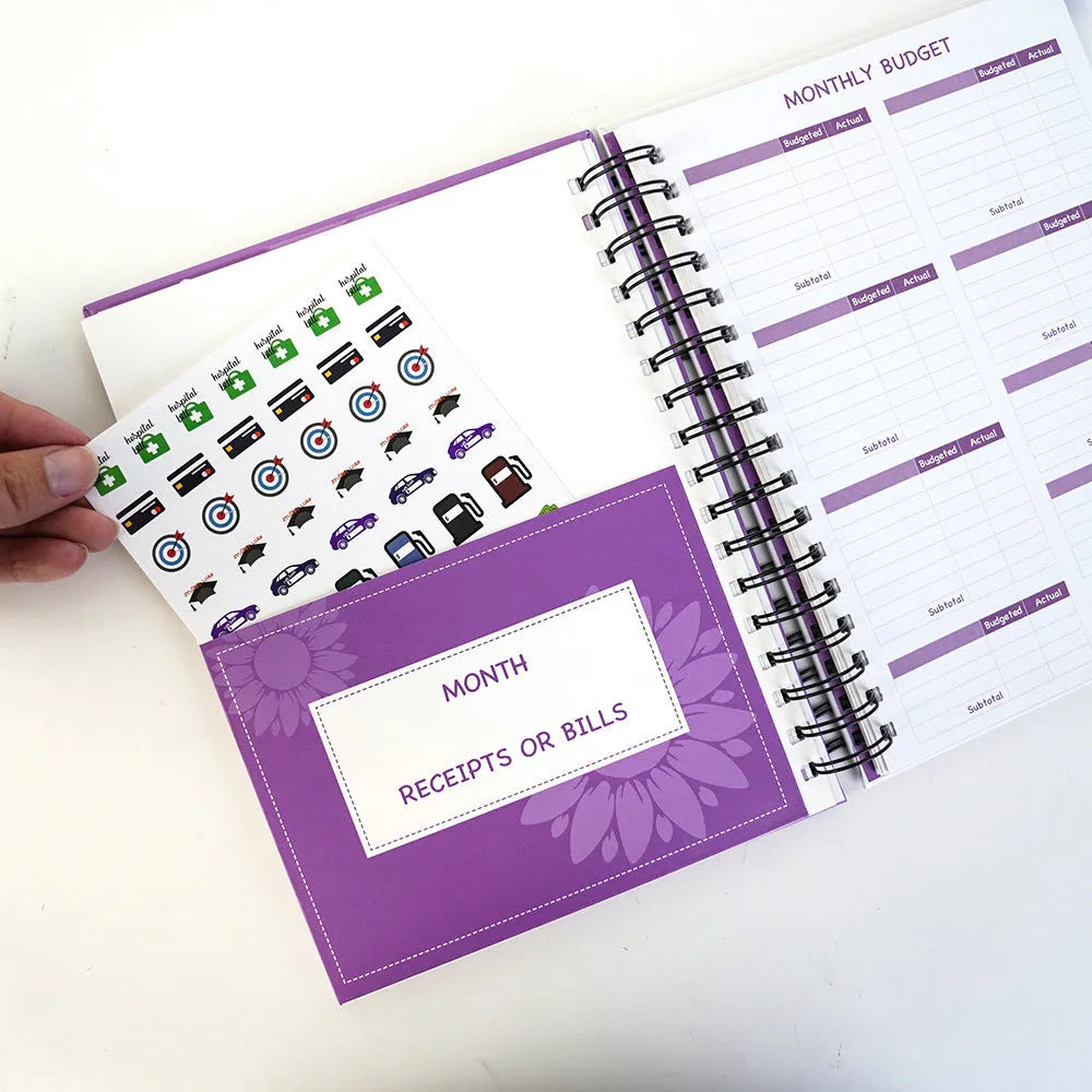 Purple coil-bound financial planning notebook open on a desk with pen, stickers, and paper clips.