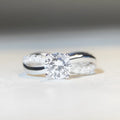 Close-up of a silver ring with a 1 karat moissanite in a four-claw setting, geometric cross design, and twisted band.