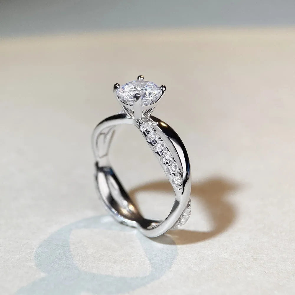 Close-up of a silver ring with a 1 karat moissanite in a four-claw setting, geometric cross design, and twisted band.