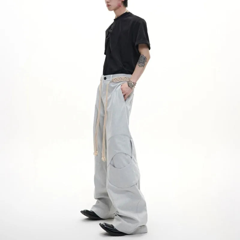 Men's dark blue loose fit straight-leg trousers showing 3D cutting, belt stitching, and tassel details on pockets and zipper.
