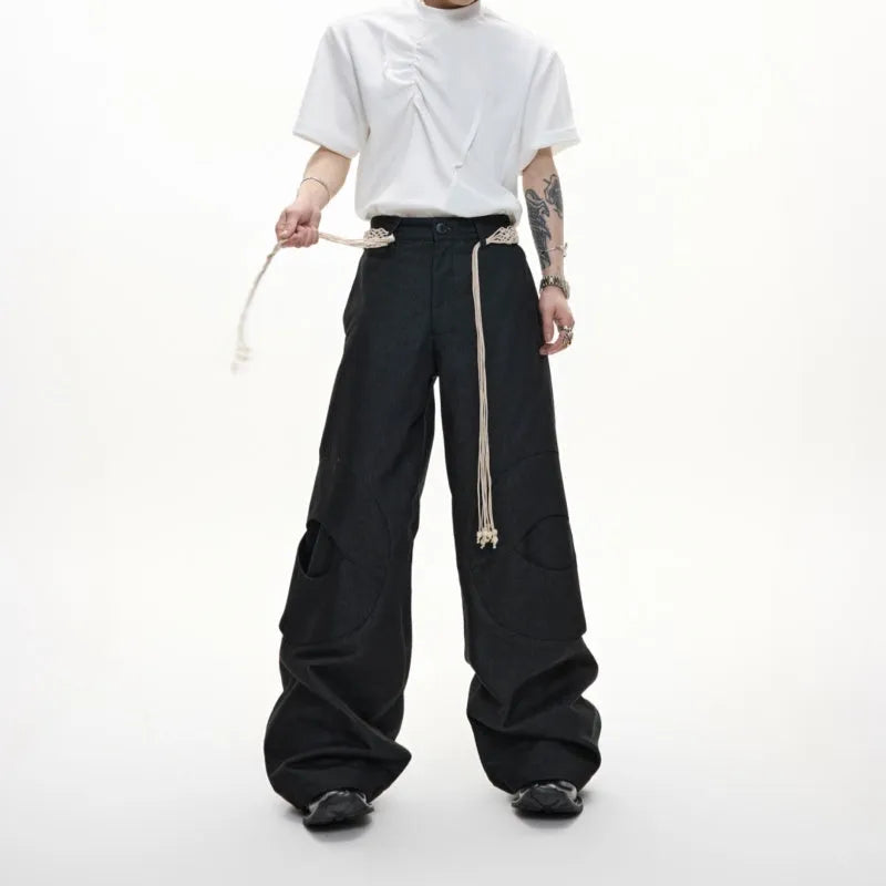 Men's dark blue loose fit straight-leg trousers showing 3D cutting, belt stitching, and tassel details on pockets and zipper.