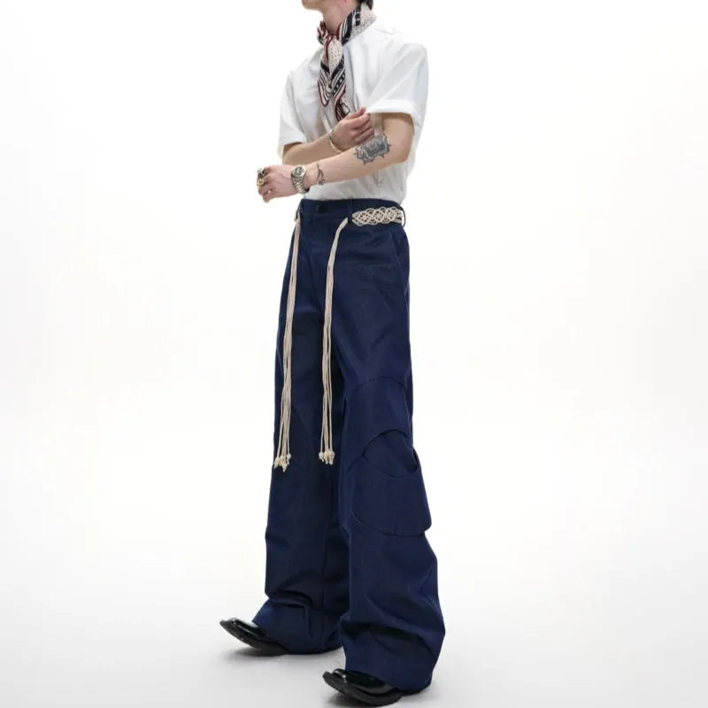 Men's dark blue loose fit straight-leg trousers showing 3D cutting, belt stitching, and tassel details on pockets and zipper.